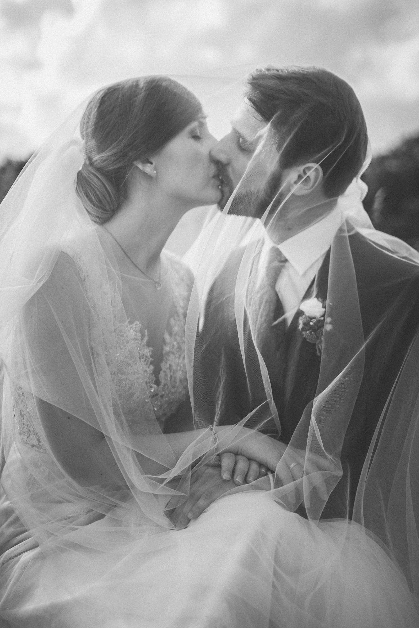 A bride and groom share a tender kiss under a veil in a beautiful black and white wedding photograph.