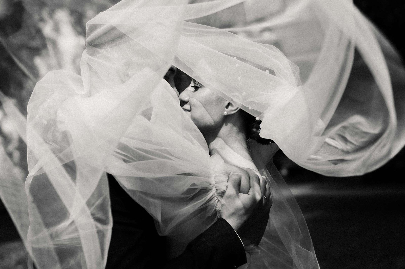 Bride and groom share a tender kiss beneath a flowing veil in elegant grayscale.