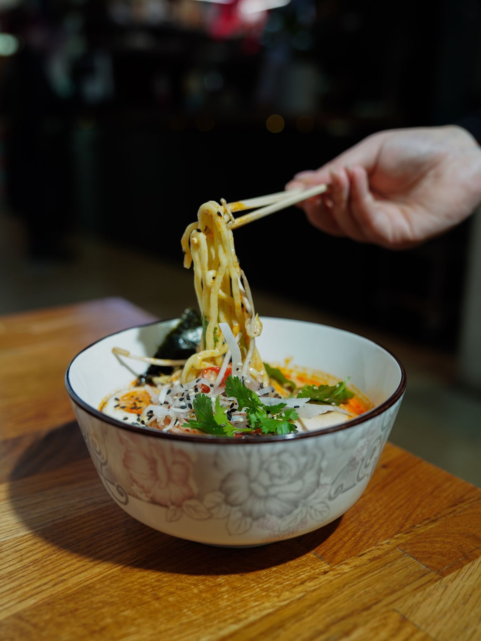 A hand using chopsticks to lift ramen noodles from a bowl in a restaurant setting.